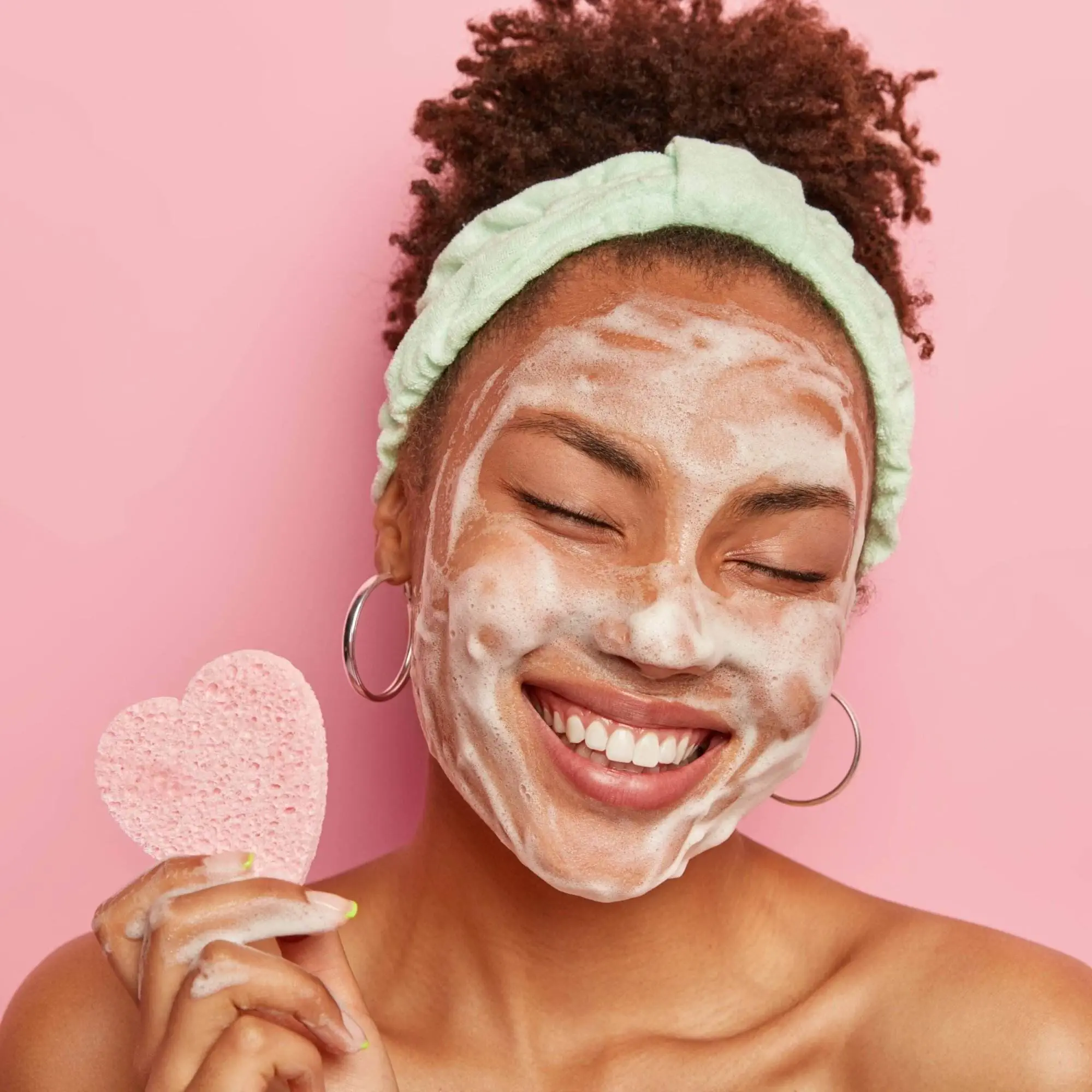 A young woman is cleansing her face with a foaming wash. She holds a pink heart-shaped sponge and wears a pink headband.
