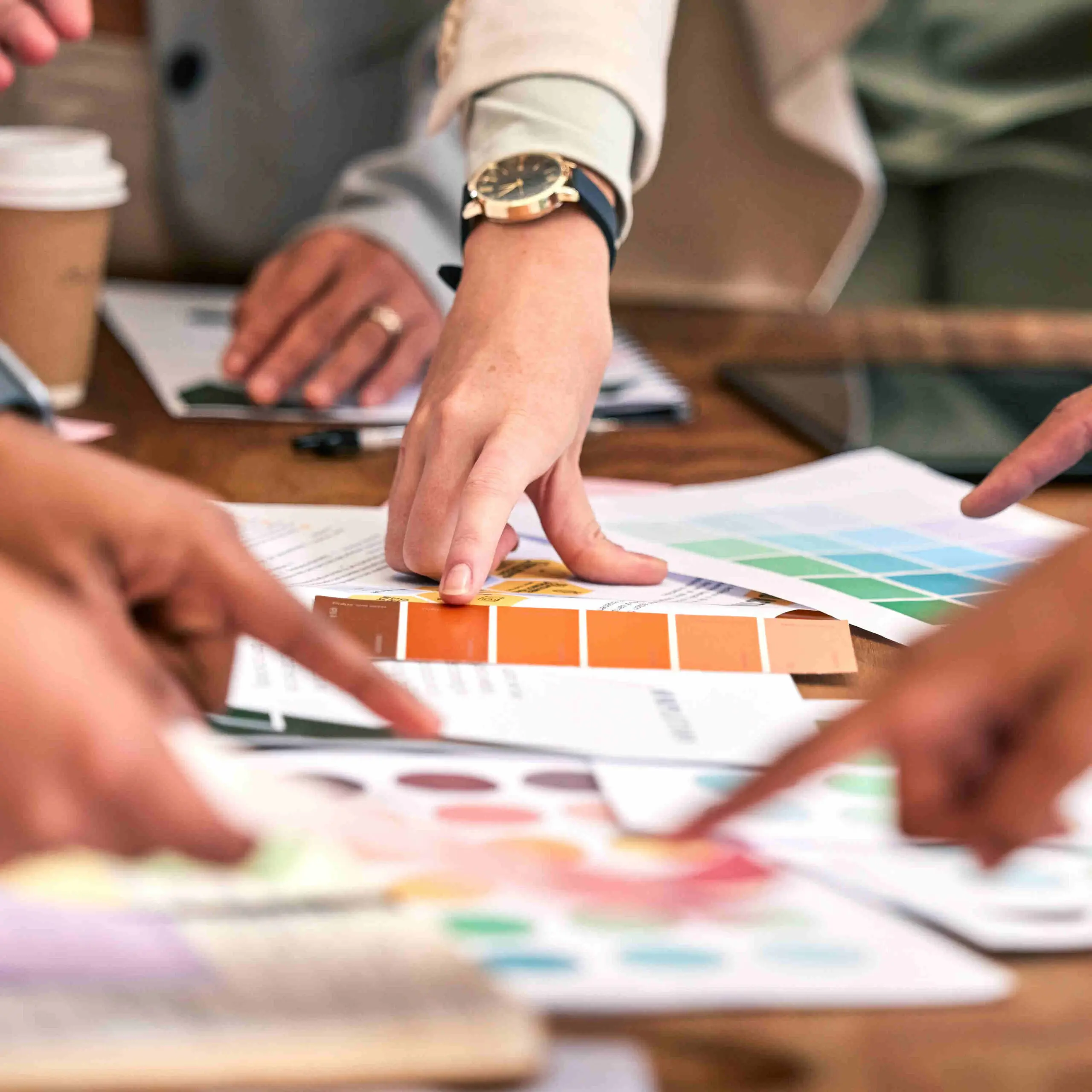 A table is filled with pages of marketing materials and branding swatches. Multiple hands are pointing at different pages.