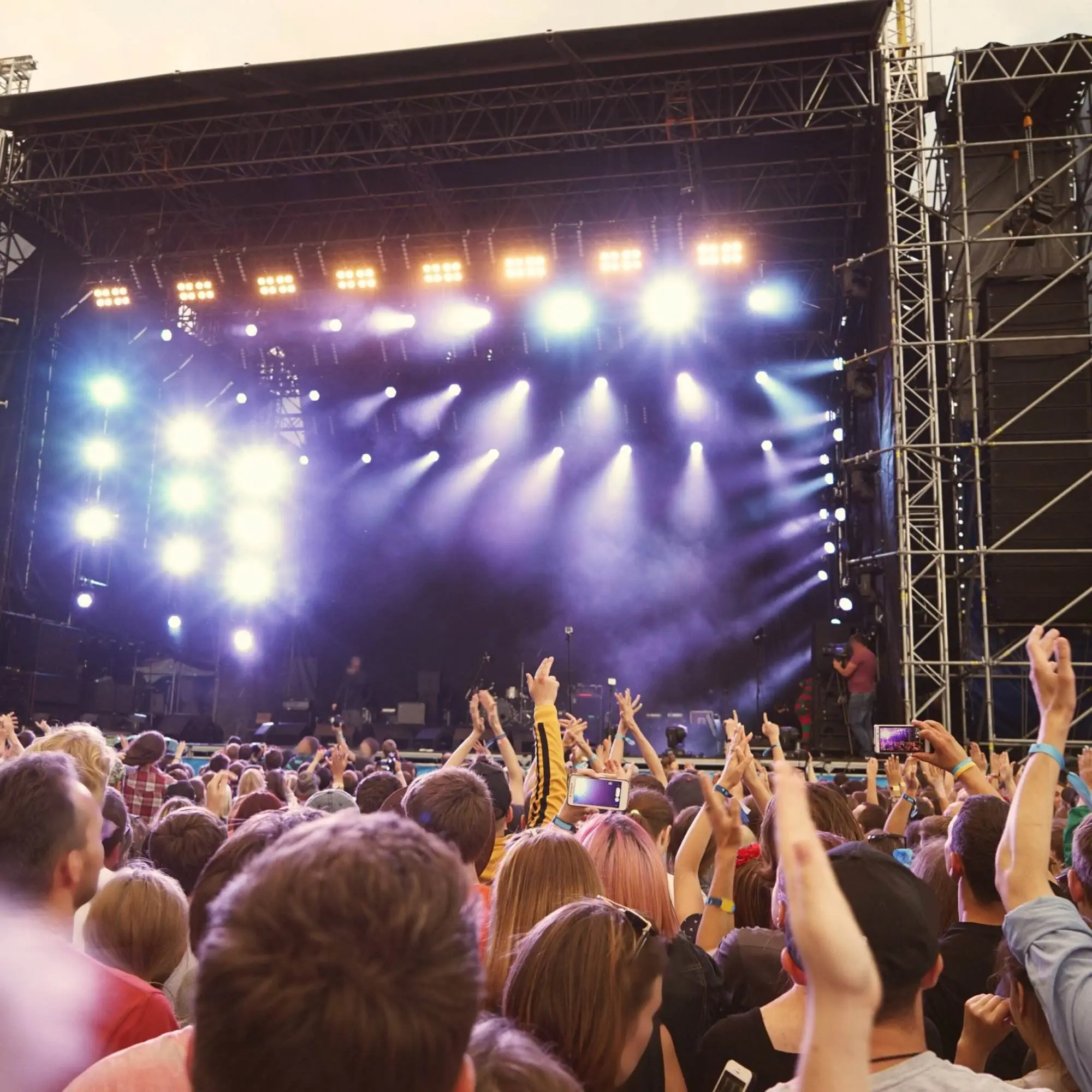 A view from within the crowd at an open-air concert during the day. A massive stage with bright lights is in the background.