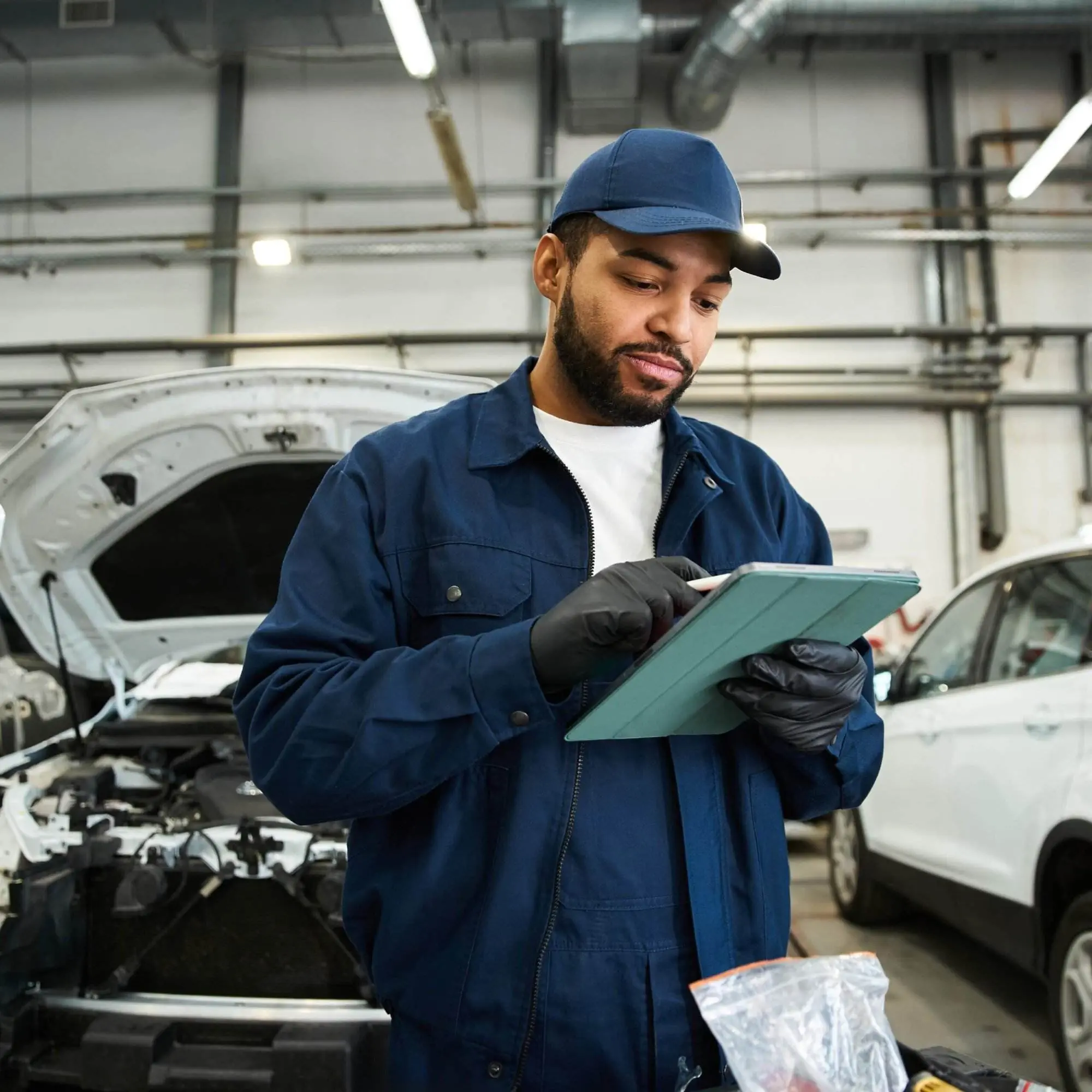 A mechanic in a blue jacket holds and looks at a tablet while standing in front of a white vehicle with the hood open.