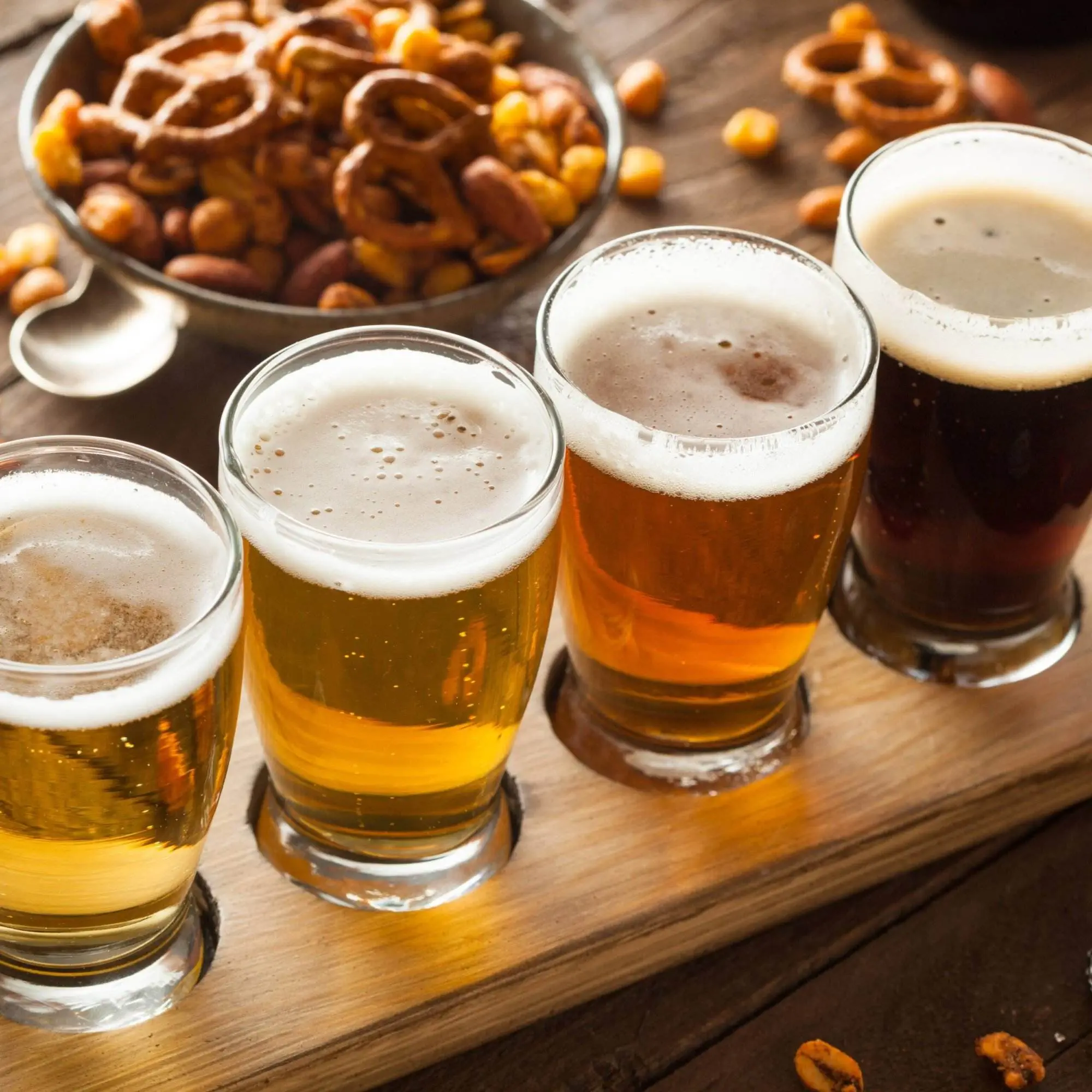 A flight of four different beers sits on a wooden paddle. A bowl of pretzels and peanuts sits next to the beers.